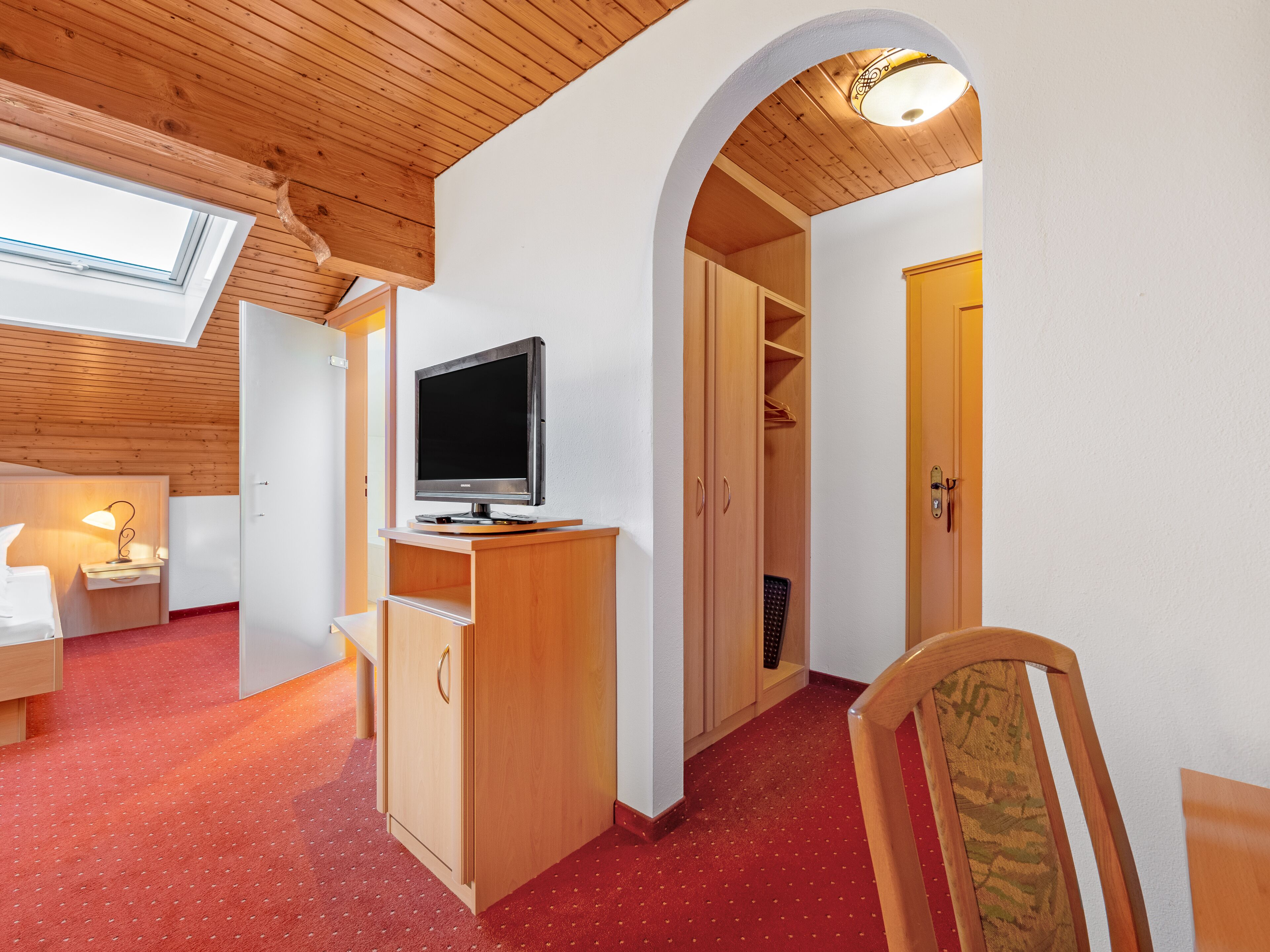 A room with a TV on a wooden stand, a desk, a chair, a wardrobe, and a red carpet.