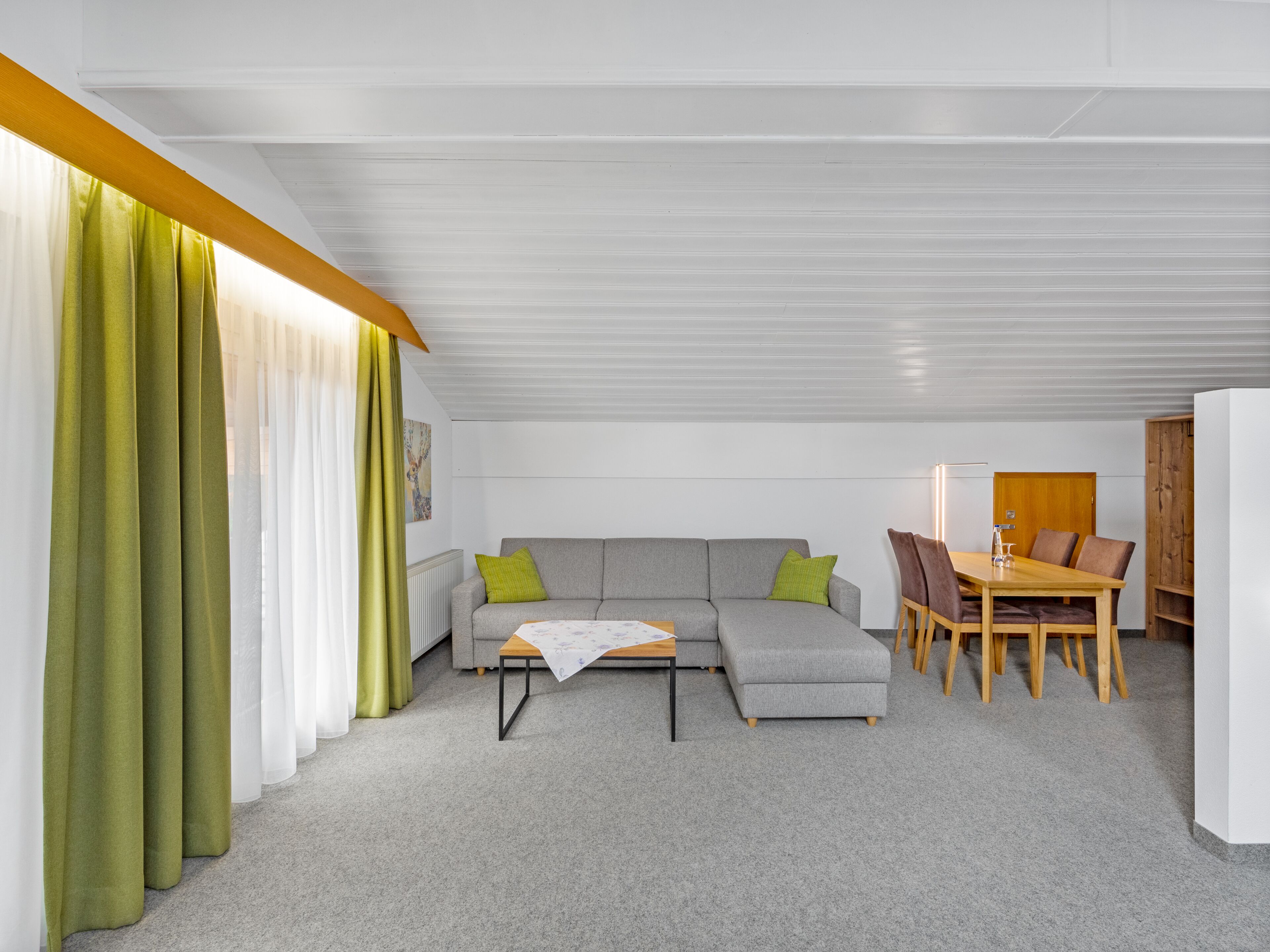 Een moderne woonkamer met een grijze bank, groene kussens, een houten salontafel en een eethoek met een houten tafel en stoelen.