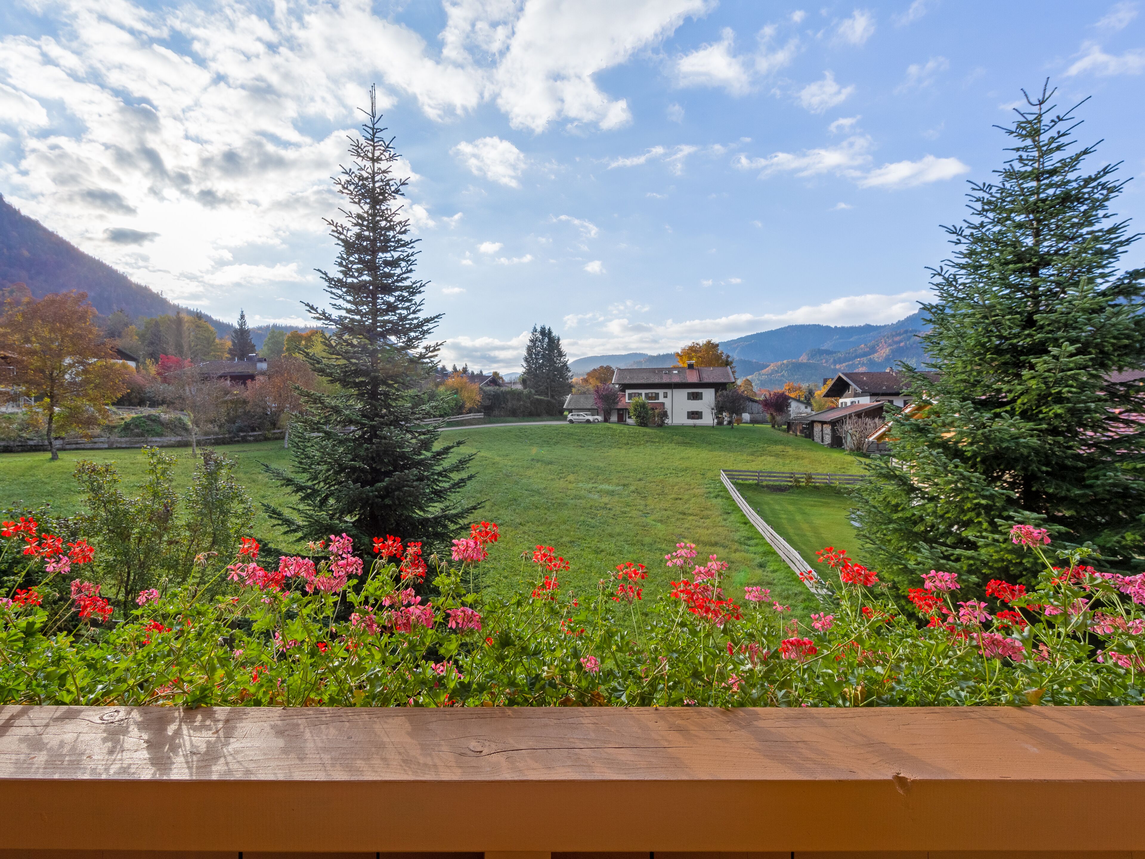 Una vista panorámica de un campo cubierto de hierba, con una terraza de madera al frente, una hilera de casas en el fondo y montañas a lo lejos.