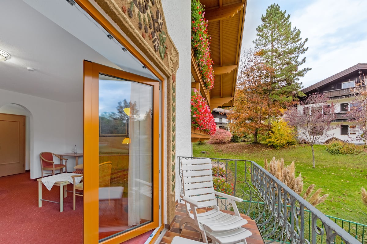 A balcony with a white chair and a table, a glass door leading to a room with a chair and a table, and a view of a garden with trees and a building in the background.