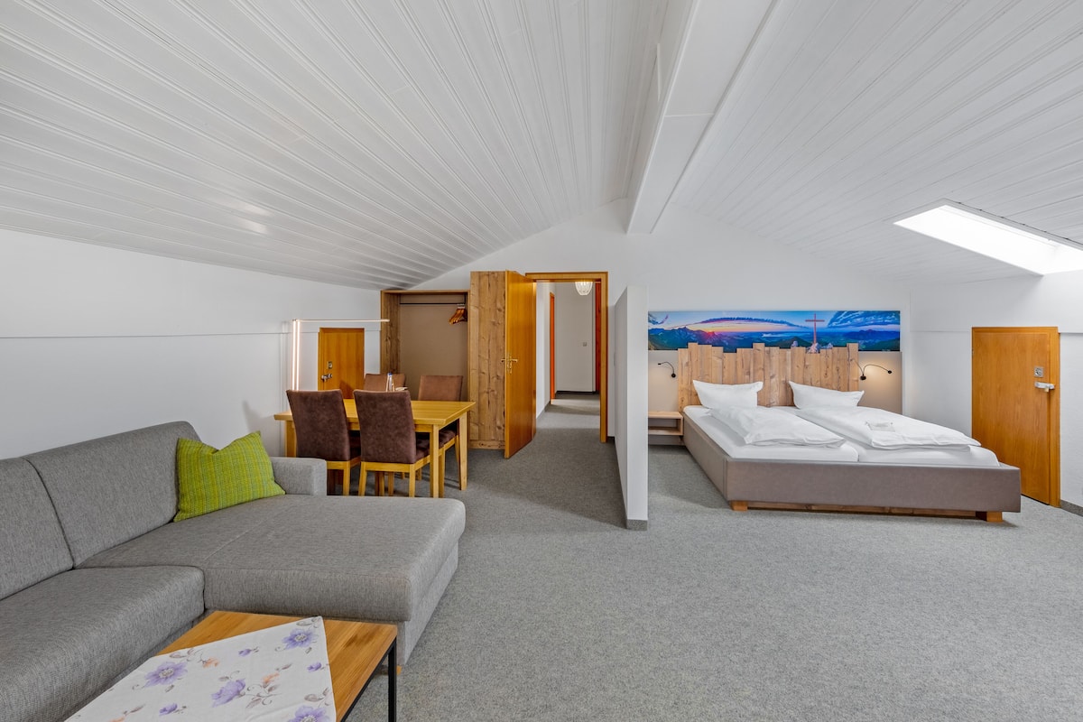 A modern hotel room with a grey sofa, a wooden dining table, a bed, and a hallway leading to another room.