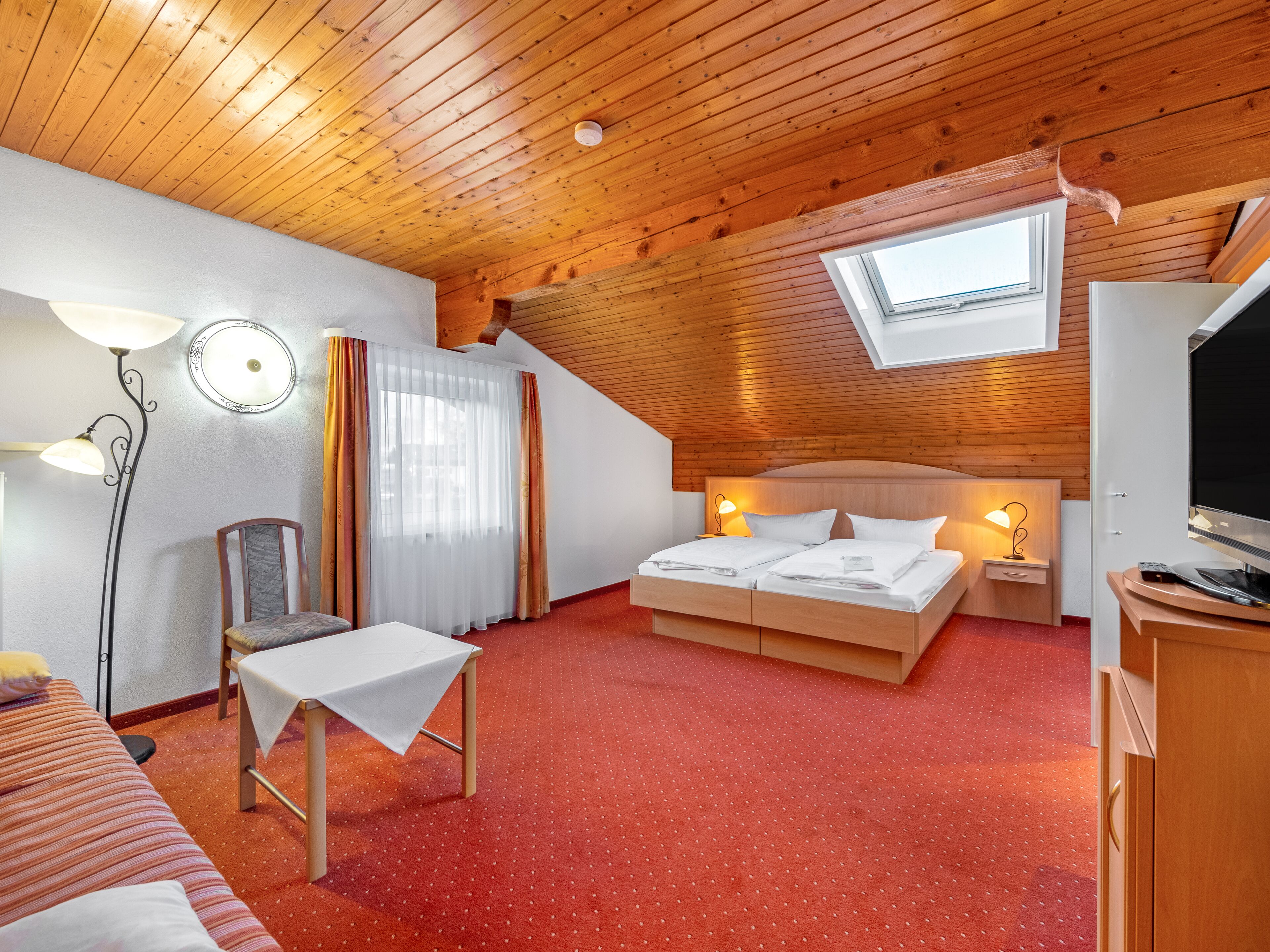 A cozy hotel room with a wooden ceiling, a large bed with two bedside tables, a chair, a small table, and a TV.