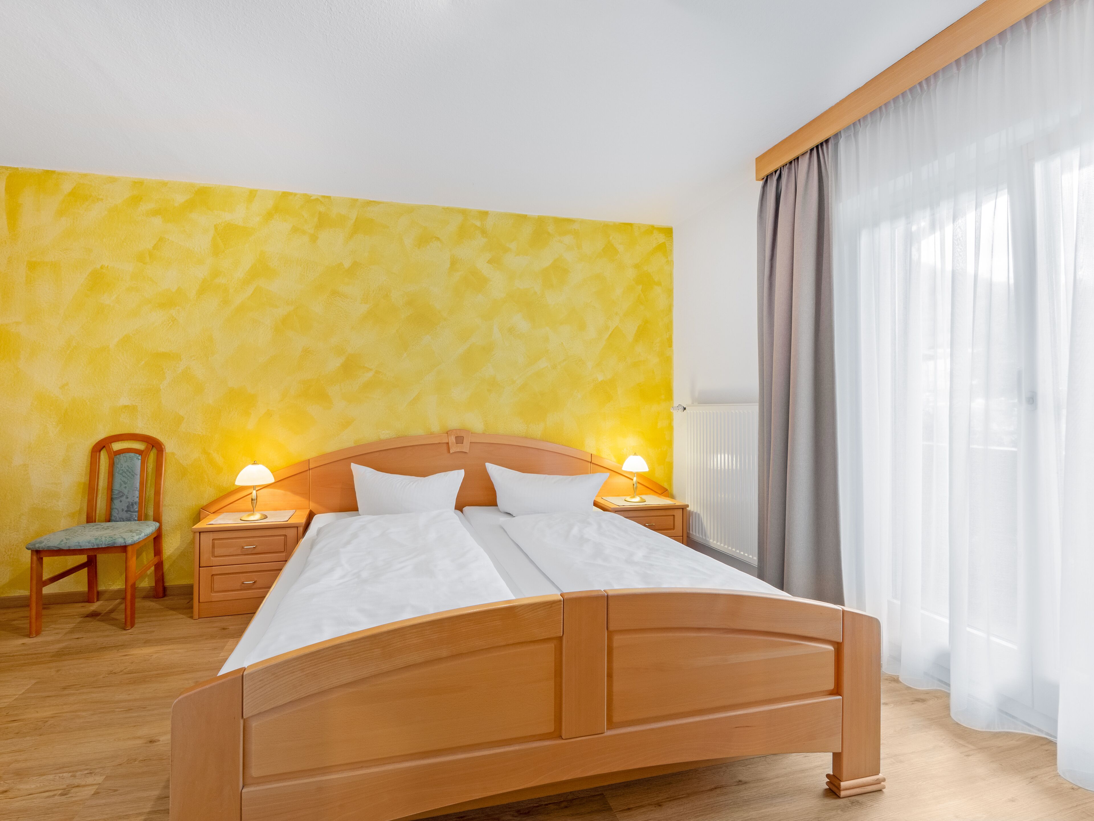 Ein Hotelzimmer mit einem hölzernen Bett, zwei Nachttischen mit Lampen, einem Sessel und einem großen Fenster mit Vorhängen.