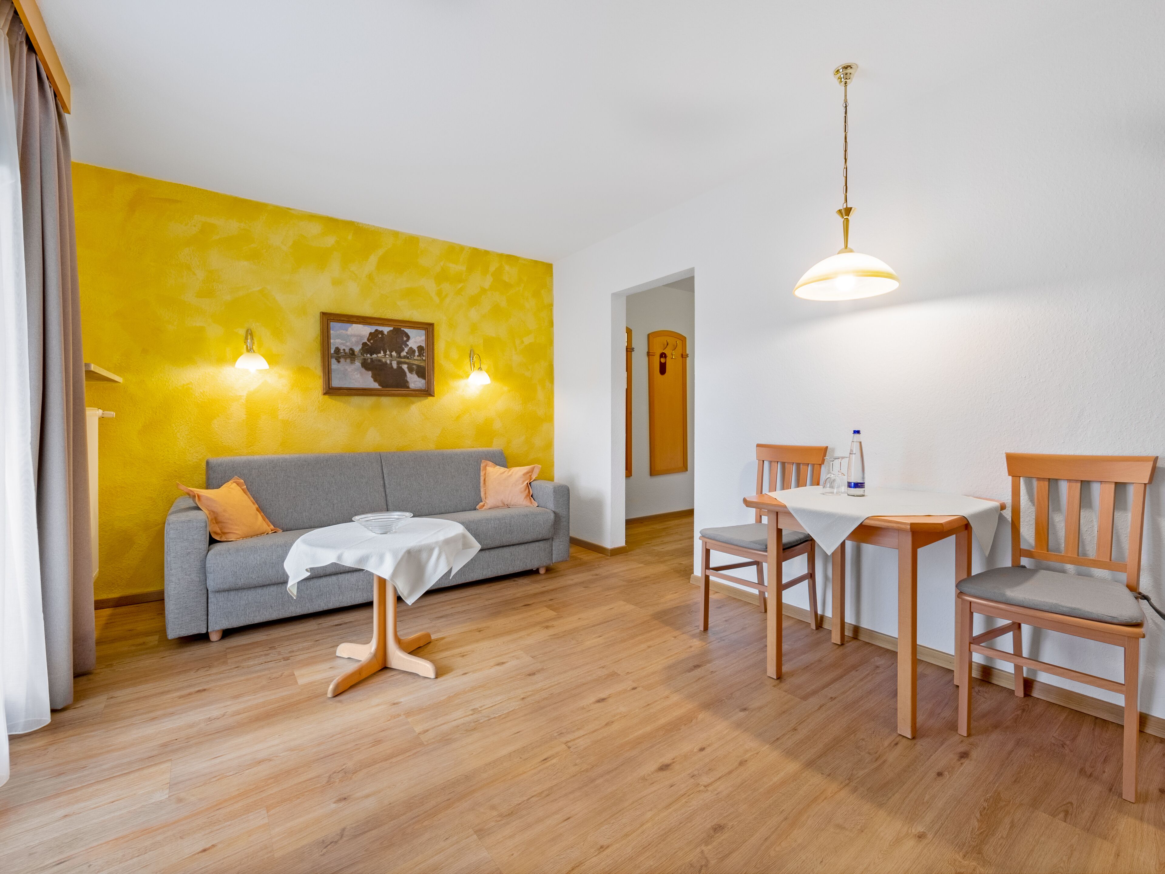 Una habitación con una pared amarilla, un sofá gris, una mesa de comedor de madera con sillas y una lámpara de techo blanca.
