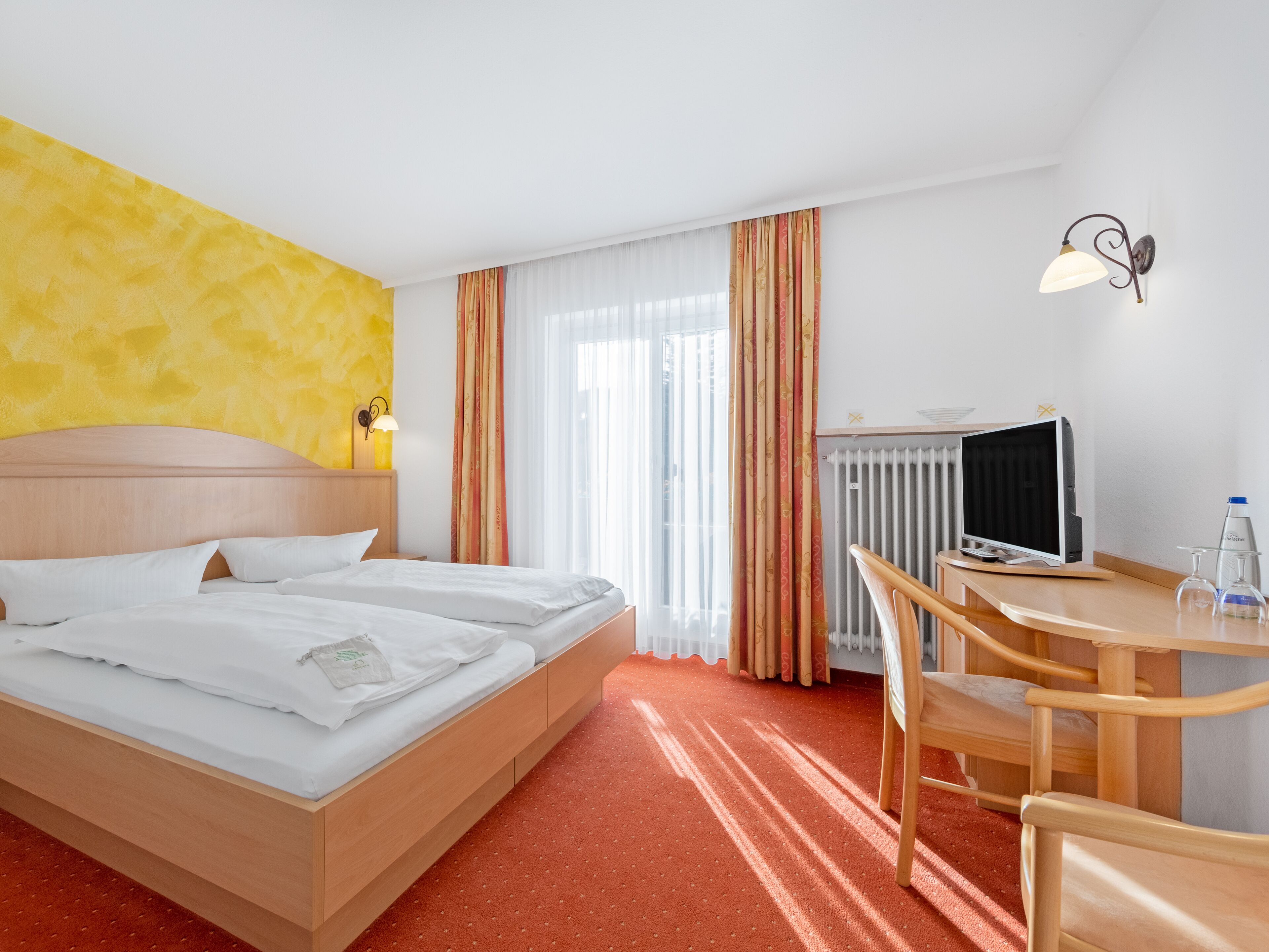 A hotel room with a bed, a desk with a computer, a chair, and a radiator.
