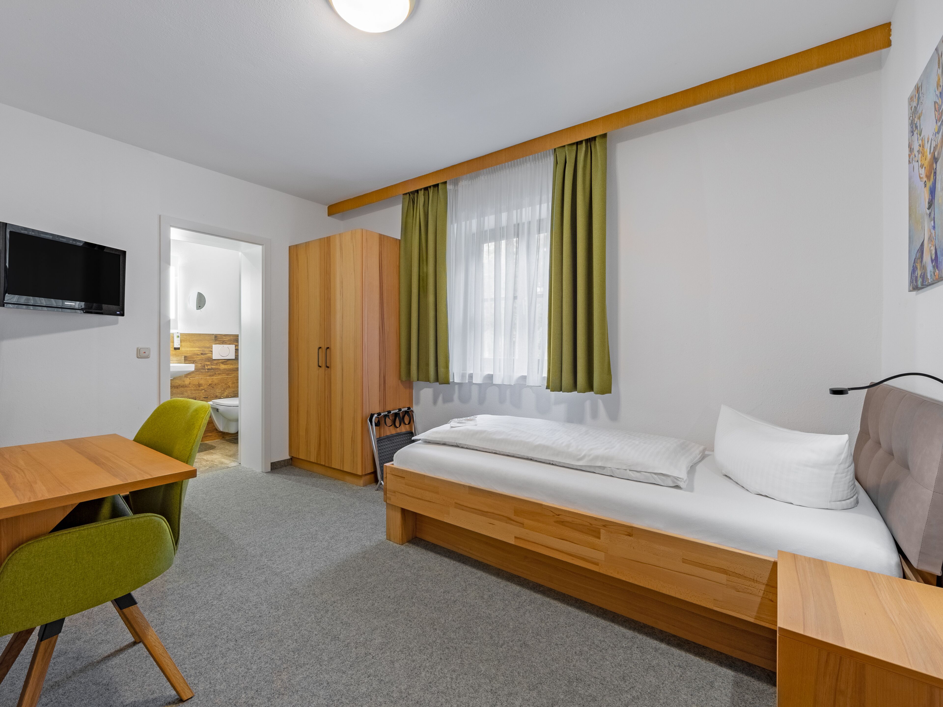 Ein kleines Hotelzimmer mit Bett, Schreibtisch, Stuhl, Kleiderschrank und Fernseher.