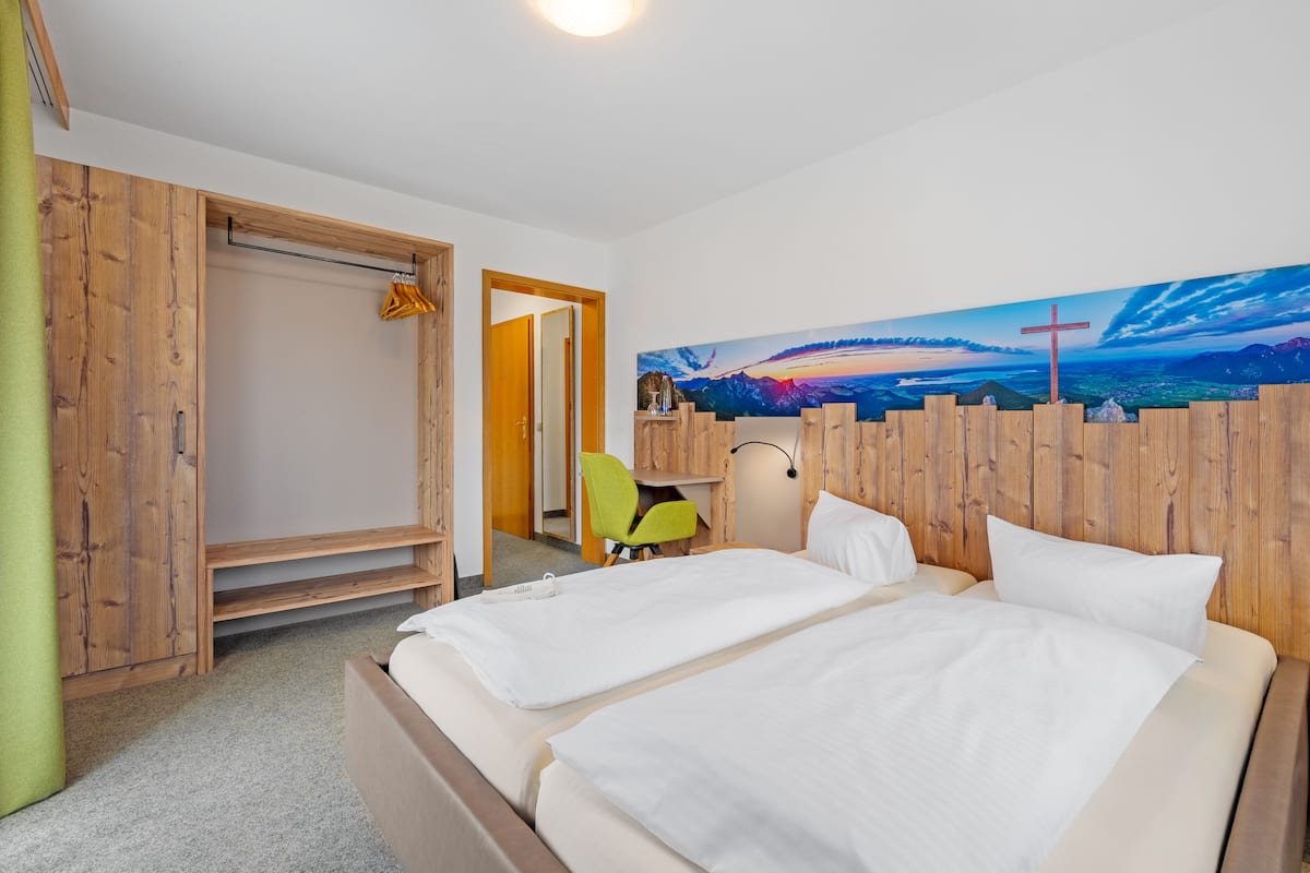 A hotel room with a large bed, a wooden wardrobe, a desk with a chair, and a painting of a landscape on the wall.