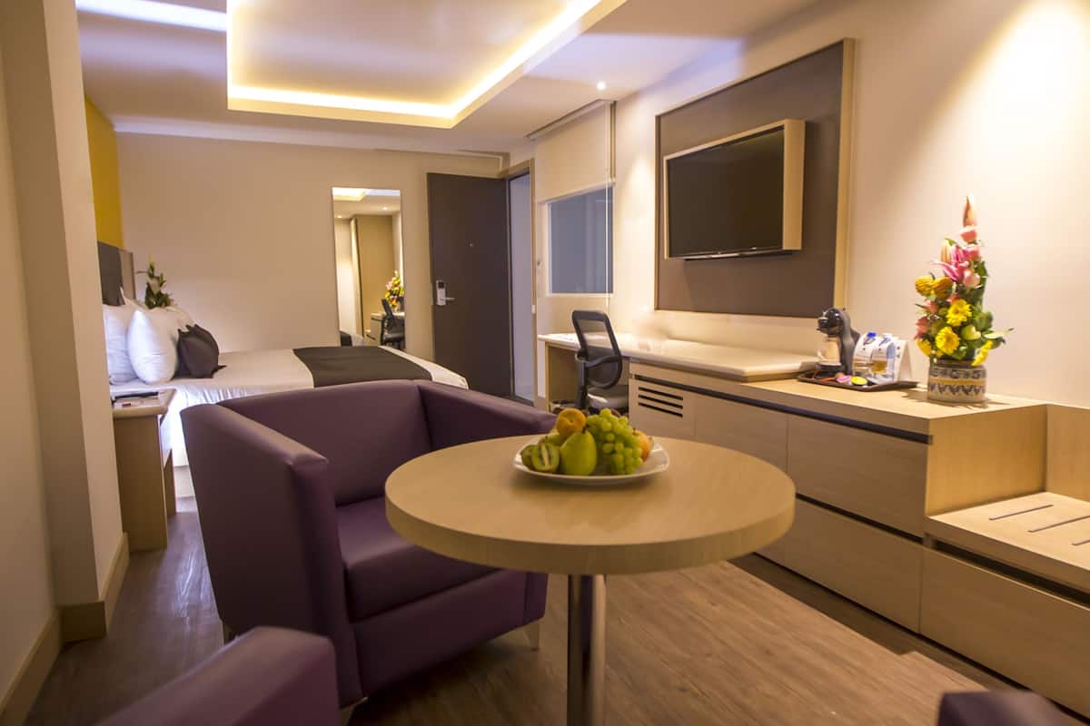 A modern hotel room with a purple armchair, a round table with fruit, a wooden bench, a TV mounted on the wall, and a kitchenette.