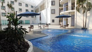 Una piscina interior con bordes de azulejos, rodeada de sillones de descanso y sombrillas, en un edificio con varios balcones y ventanas amplias.