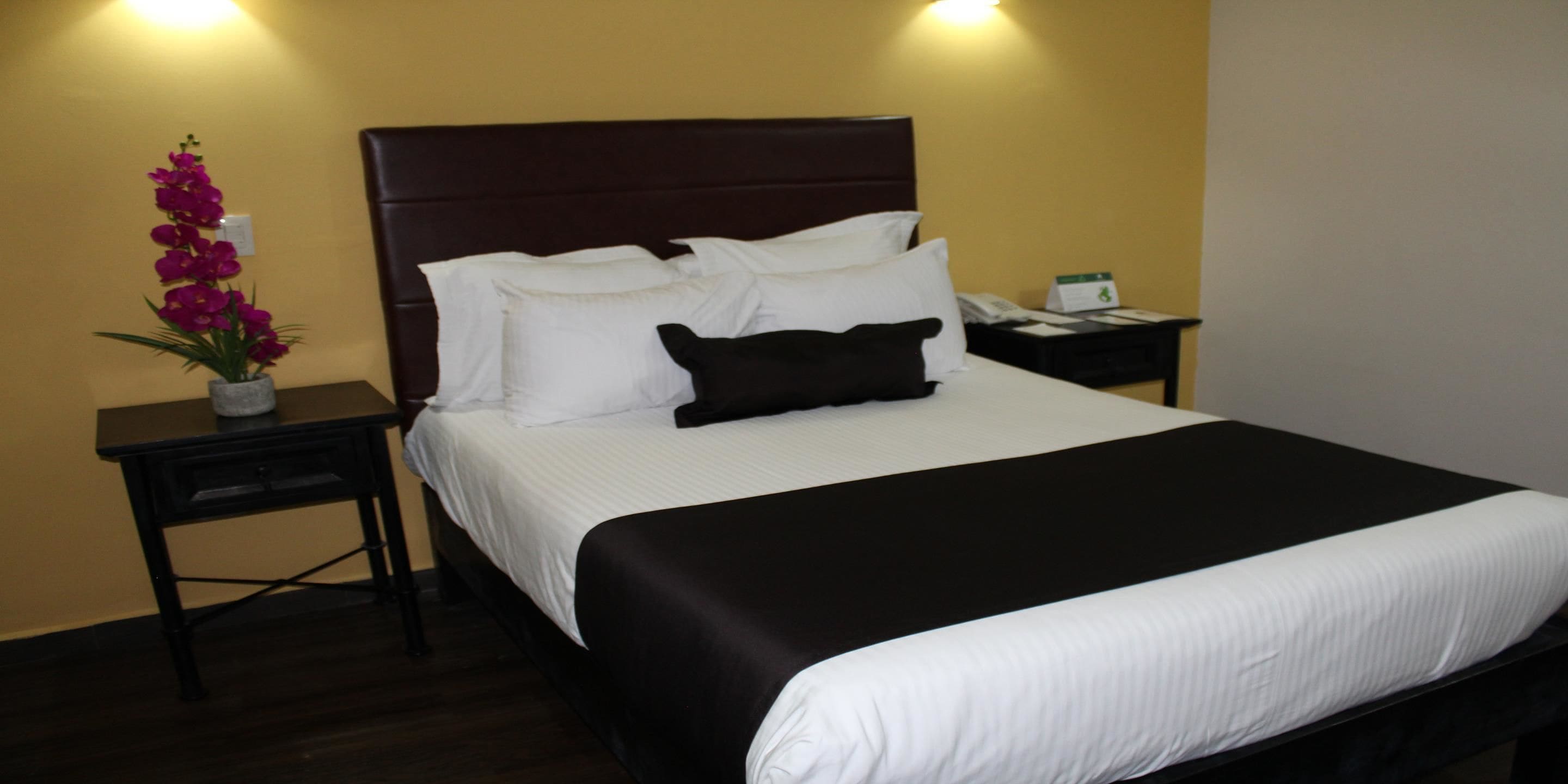 Una cama bien hecha con ropa de cama blanca y negra, un cabecero de madera oscura y una mesita de noche con una lámpara y un teléfono.