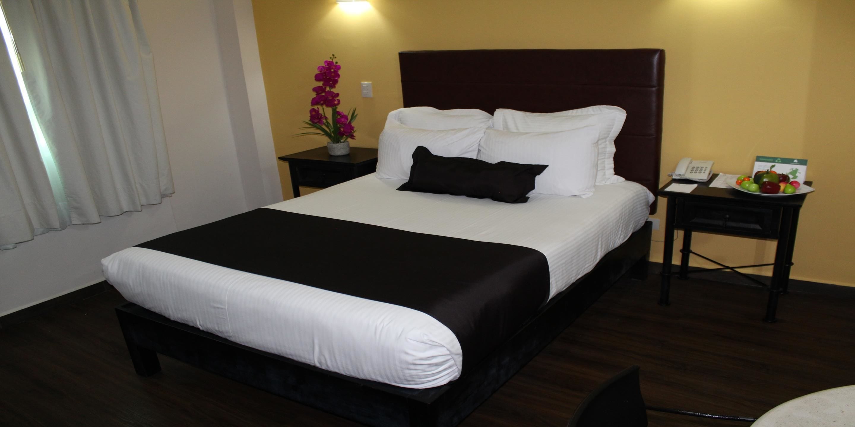 A hotel room with a large bed, a bedside table with a phone and a fruit basket, a small table with a vase of flowers, and a window with curtains.