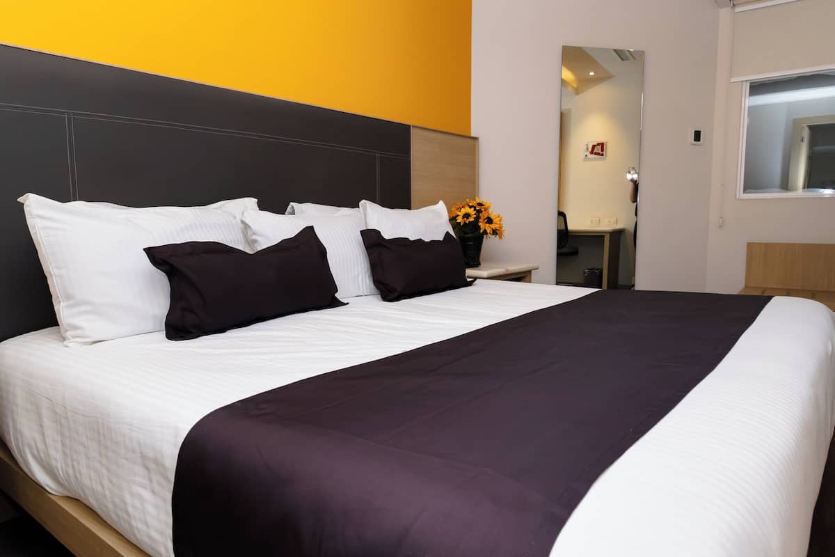 A neatly made bed with white and dark pillows, a purple bedspread, and a small table with a vase of sunflowers.