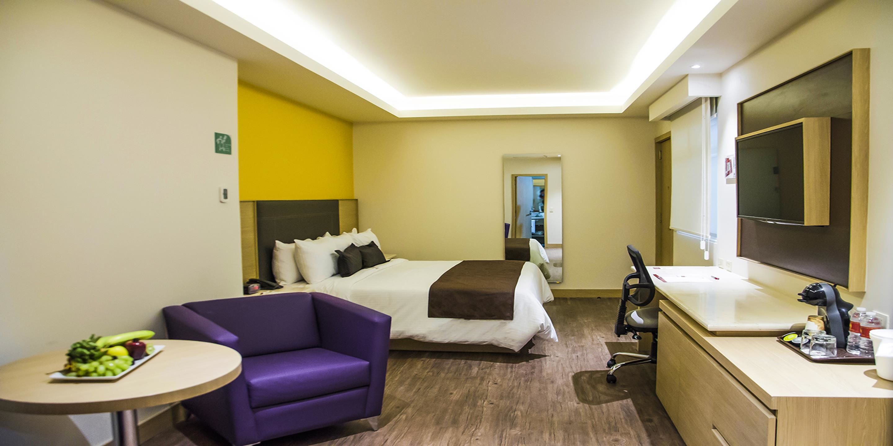 A hotel room with a bed, a purple armchair, a small round table with fruit, a desk with a chair, and a TV.