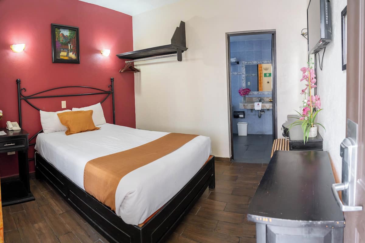 A hotel room with a bed, a nightstand, a desk, a mirror, and a bathroom visible through an open door.