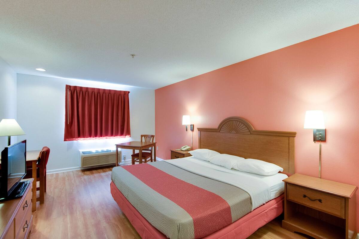 A hotel room with a bed, bedside table, lamp, desk, chair, and a television.