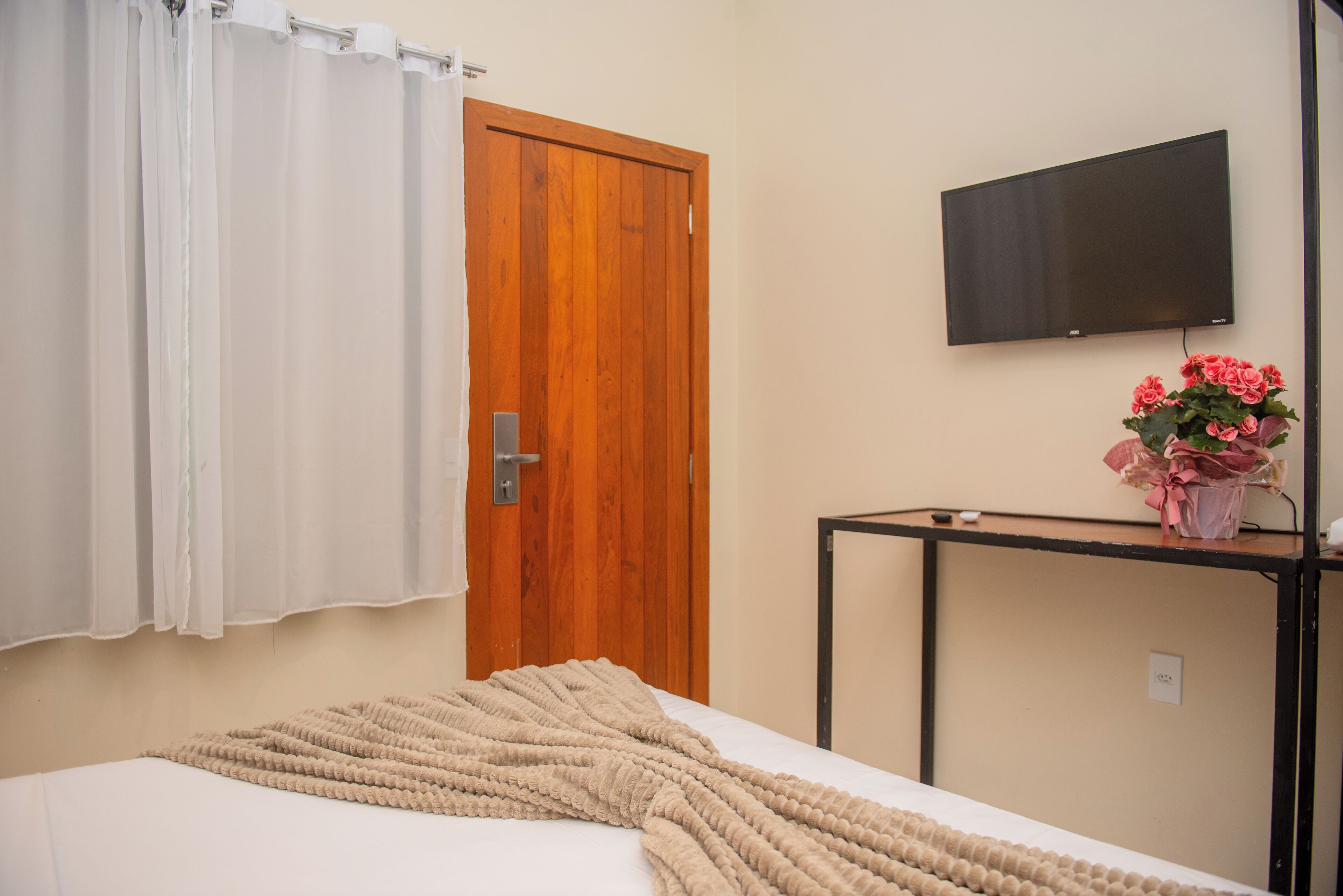 Quarto com cama, porta de madeira, televisão na parede e pequena mesa com um vaso de flores.