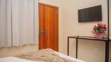 Quarto com cama, porta de madeira, televisão na parede e pequena mesa com um vaso de flores.