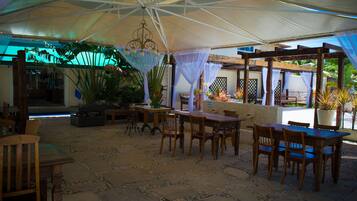 An outdoor patio area with wooden tables and chairs, a large umbrella providing shade, and potted plants.