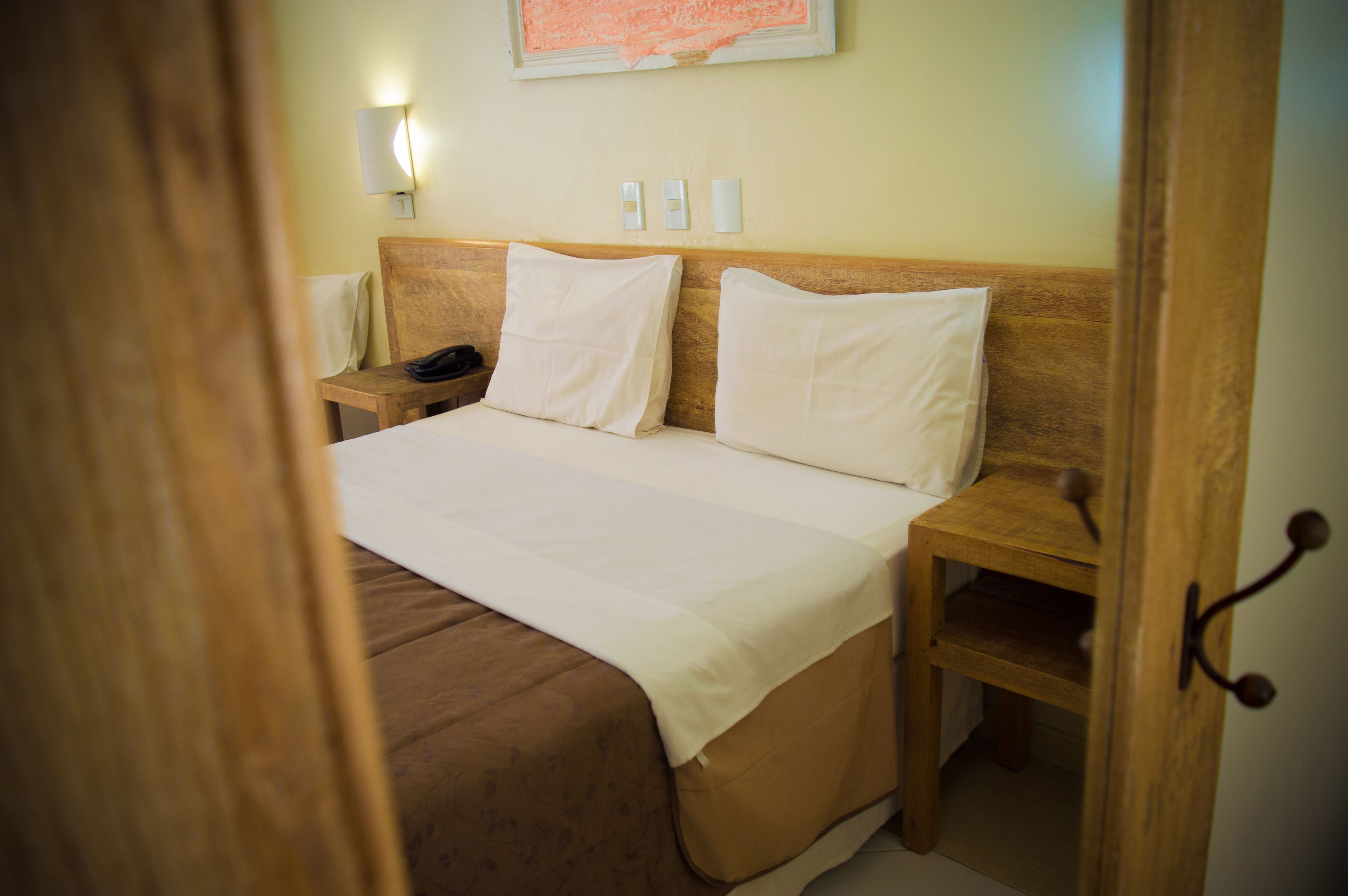 A hotel room with a wooden headboard, a bed with white linens, a bedside table with a lamp, and a wall-mounted light fixture.