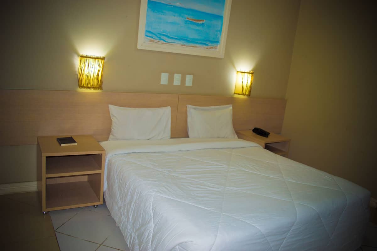 Una cama bien hecha con ropa blanca, dos mesitas de noche con lámparas y un cuadro de una playa en la pared.