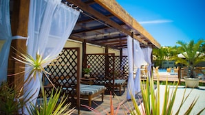 A covered outdoor seating area with wooden furniture and a pool in the background. - Don Quijote Hotel (Búzios)