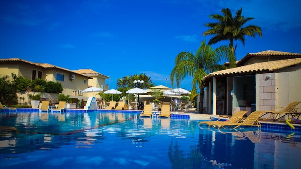 A swimming pool with lounge chairs and umbrellas, surrounded by palm trees and buildings with tiled roofs. - Don Quijote Hotel (Búzios)