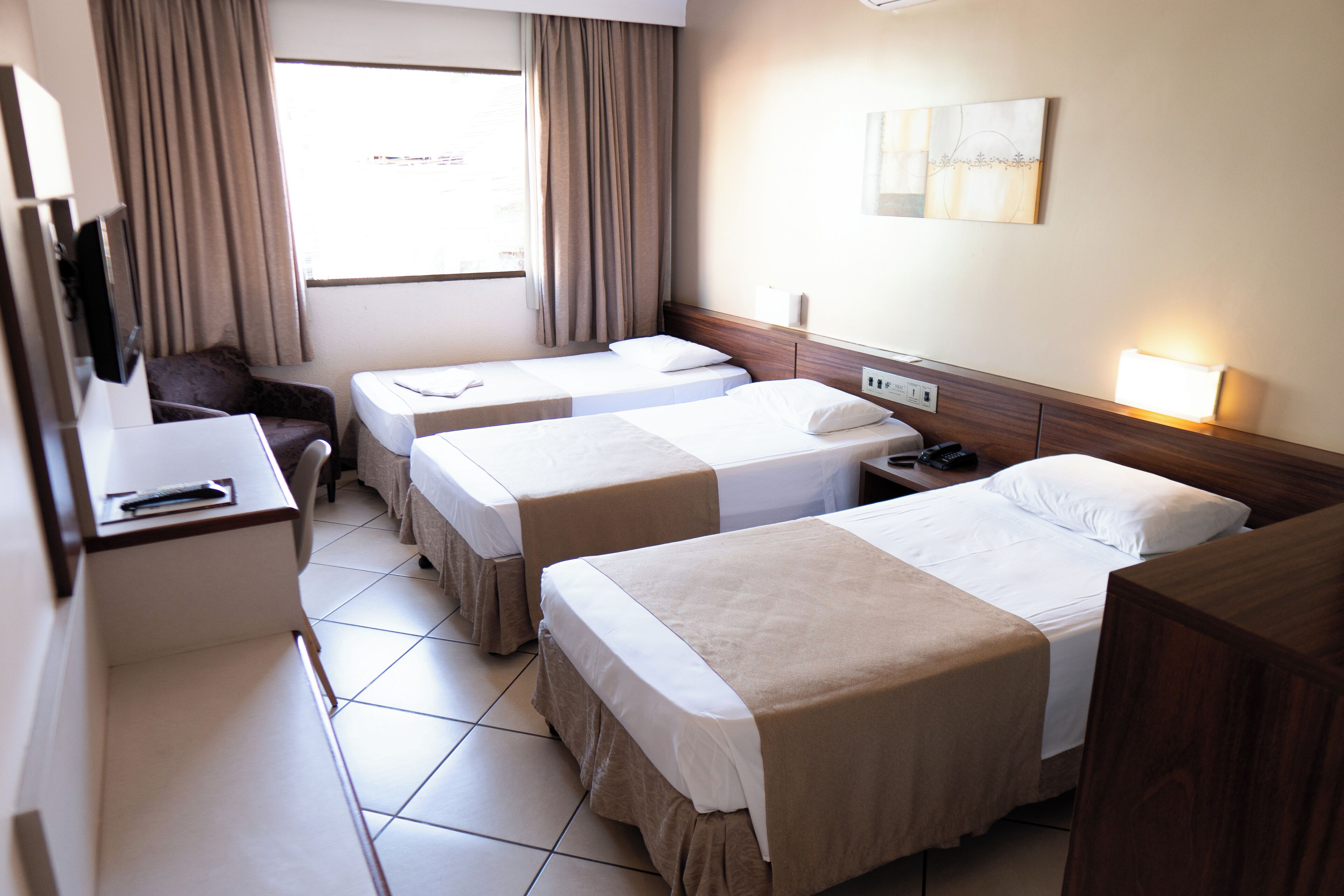 Habitación de hotel con dos camas individuales, un escritorio con silla, televisor y ventana con cortinas.