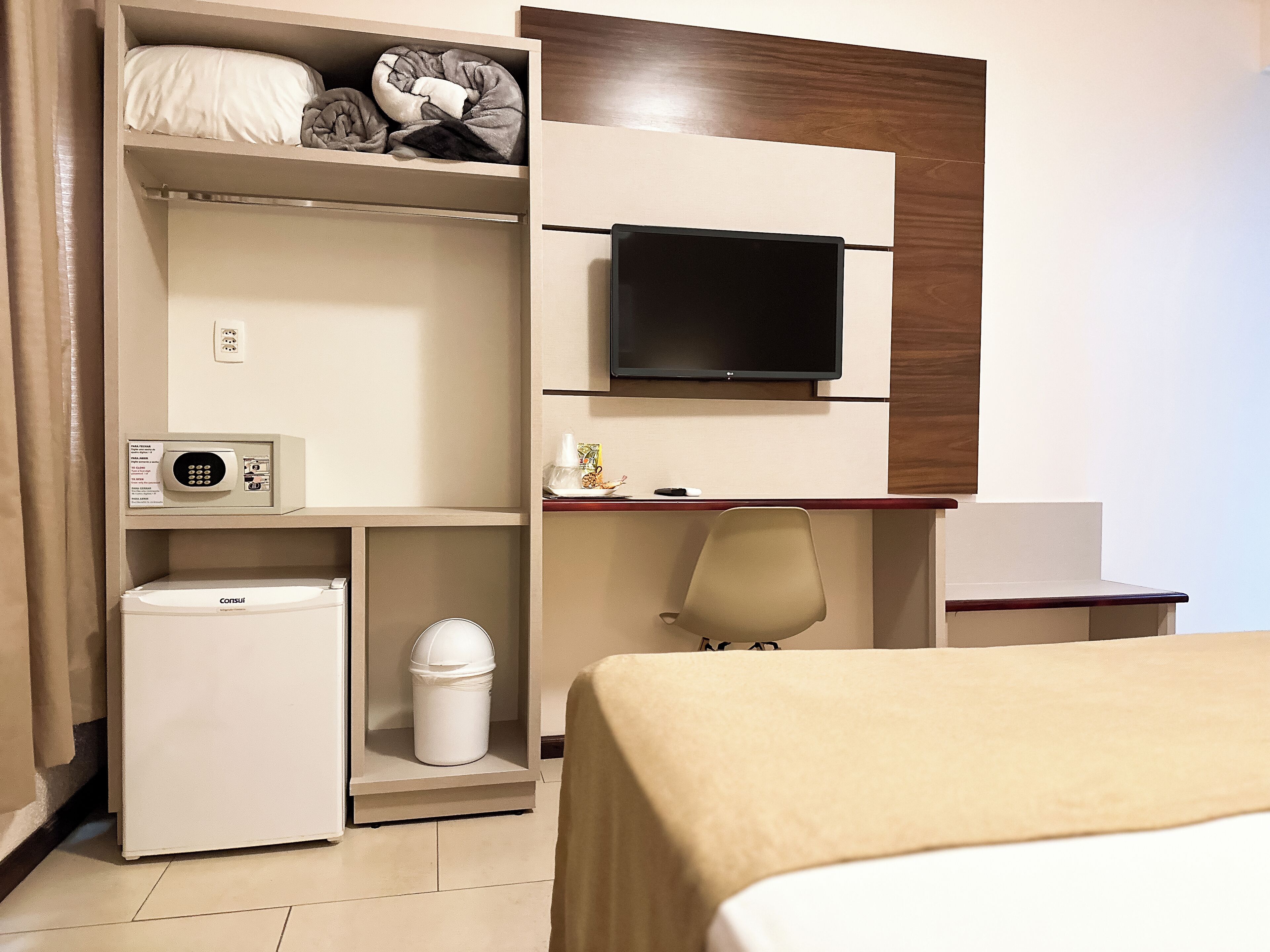 Een hotelkamer met een bed, een aan de muur bevestigde televisie, een minibar, een prullenbak en een bureau met een stoel.