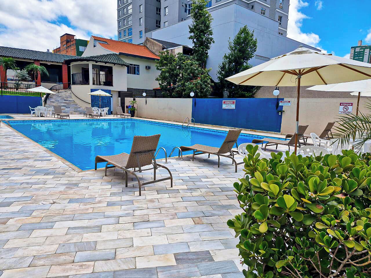 Piscina com espreguiçadeiras e guarda-sóis, cercada por um deck ladrilhado e vegetação.