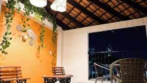 An outdoor seating area with wooden benches and wicker chairs, a tiled floor, and a view of the city at night. - Hotel San Rafael Comfort Class (Foz do Iguaçu)