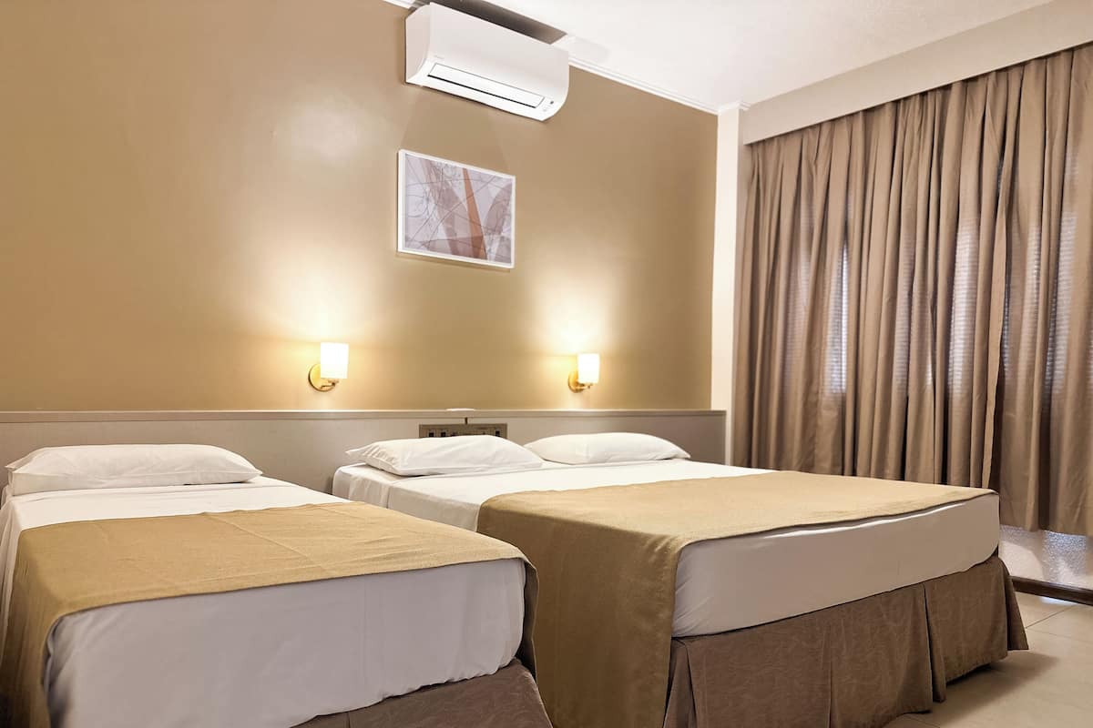 Une chambre d’hôtel avec deux lits, des murs de couleur beige, un ventilateur de plafond et une fenêtre avec des rideaux.