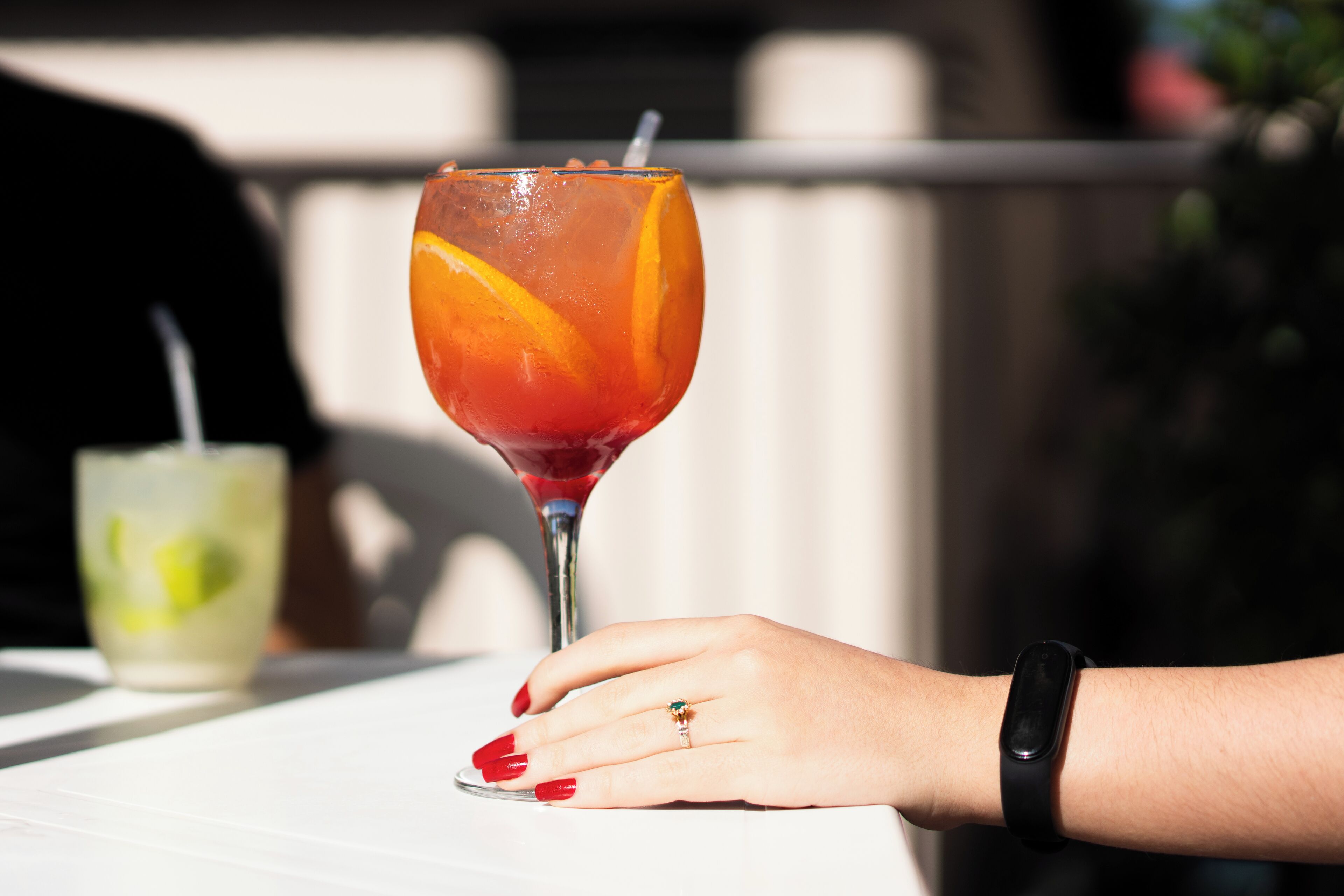 Une main portant une montre connectée, posée sur une table, à côté d’un verre contenant une boisson rouge et une tranche d’orange.