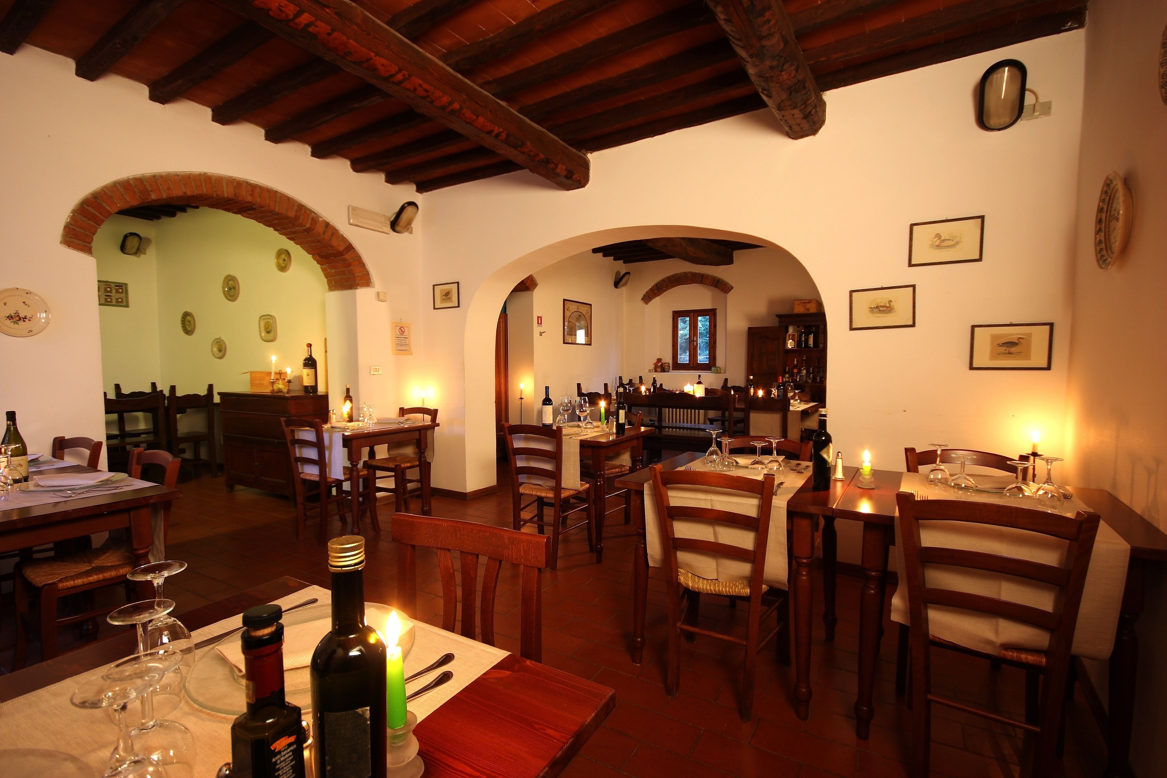 A restaurant with wooden tables and chairs, wine bottles, and candles. There are arches and a bar area in the background.