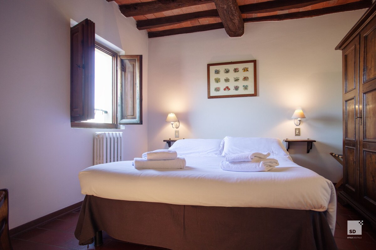 A neatly made bed with white linens and towels, a wooden headboard, a window with a view, and a wall-mounted picture frame.