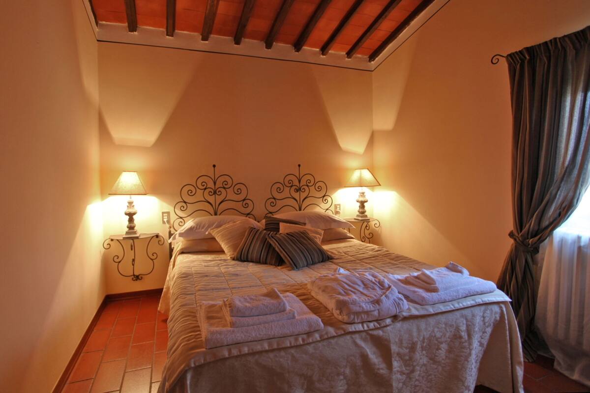 Una cama bien hecha con almohadas y una colcha, dos lámparas de noche y un ventanal con cortinas.