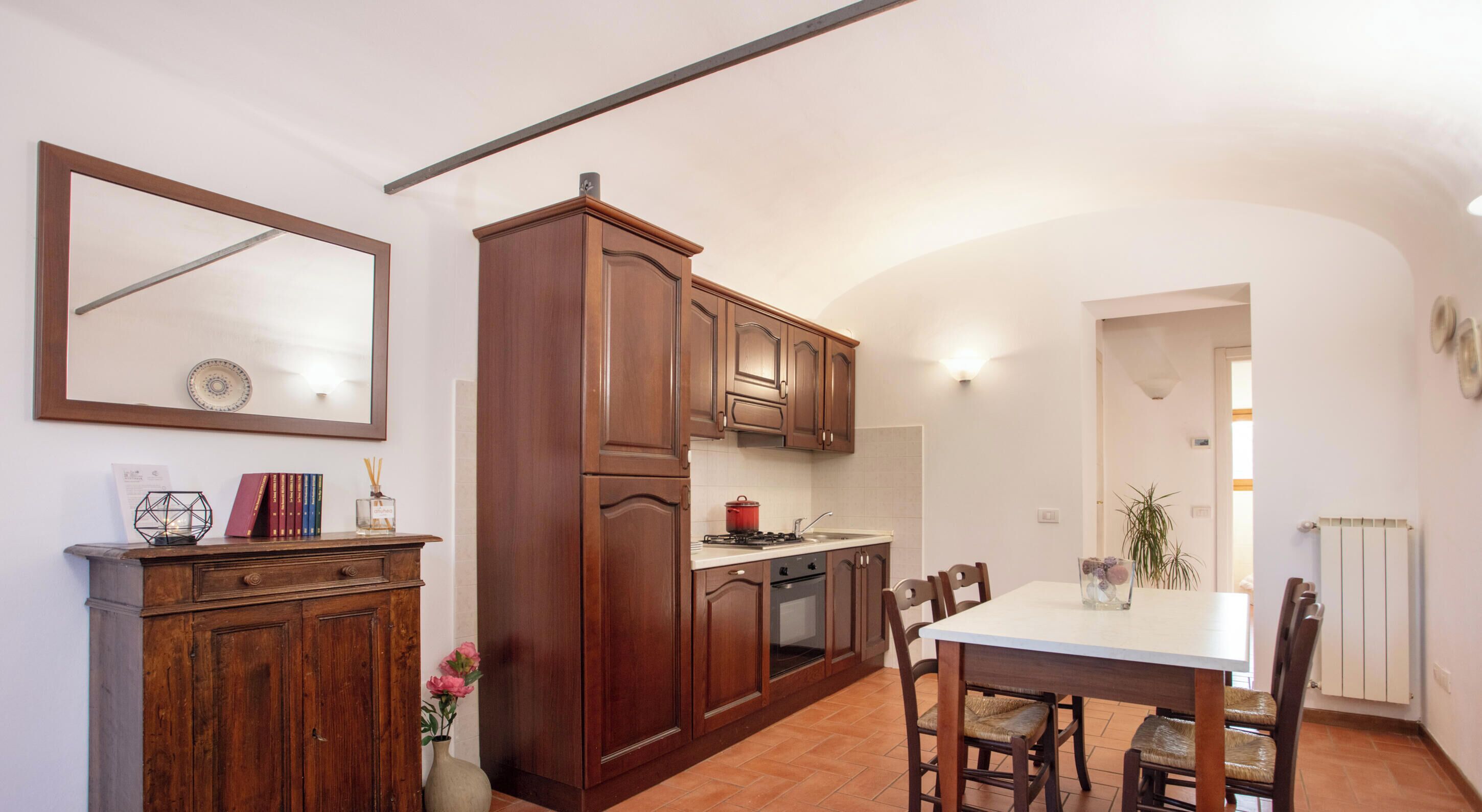 Een keuken met houten kastjes, een eettafel, stoelen, een fornuis en een spiegel aan de muur.