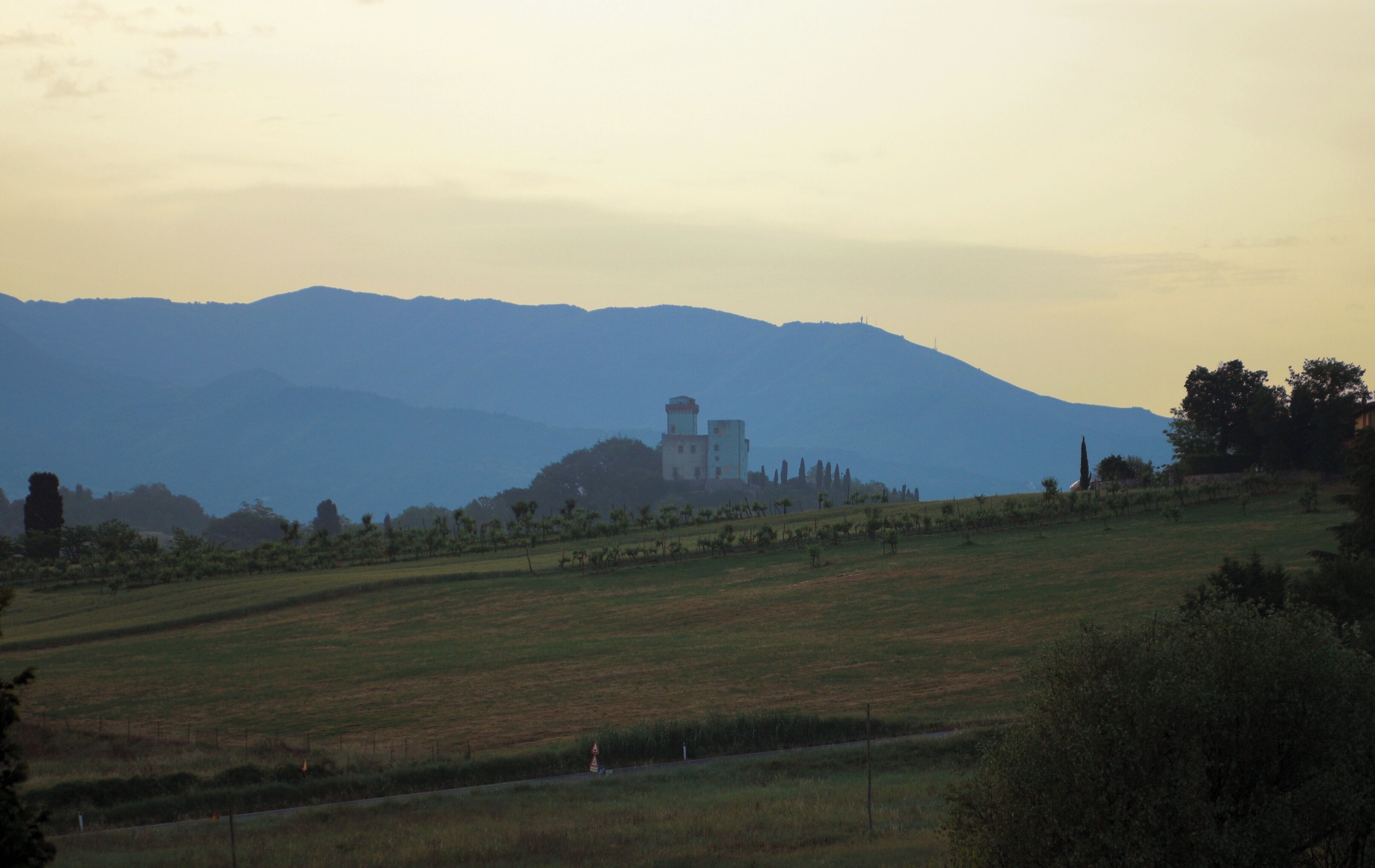A rural landscape with a prominent building on a hill, surrounded by fields and distant mountains.