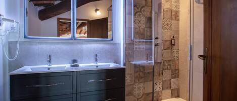 Une salle de bain moderne avec deux vasques, des meubles de rangement aux couleurs sombres, un grand miroir et une douche séparée par une paroi de verre.