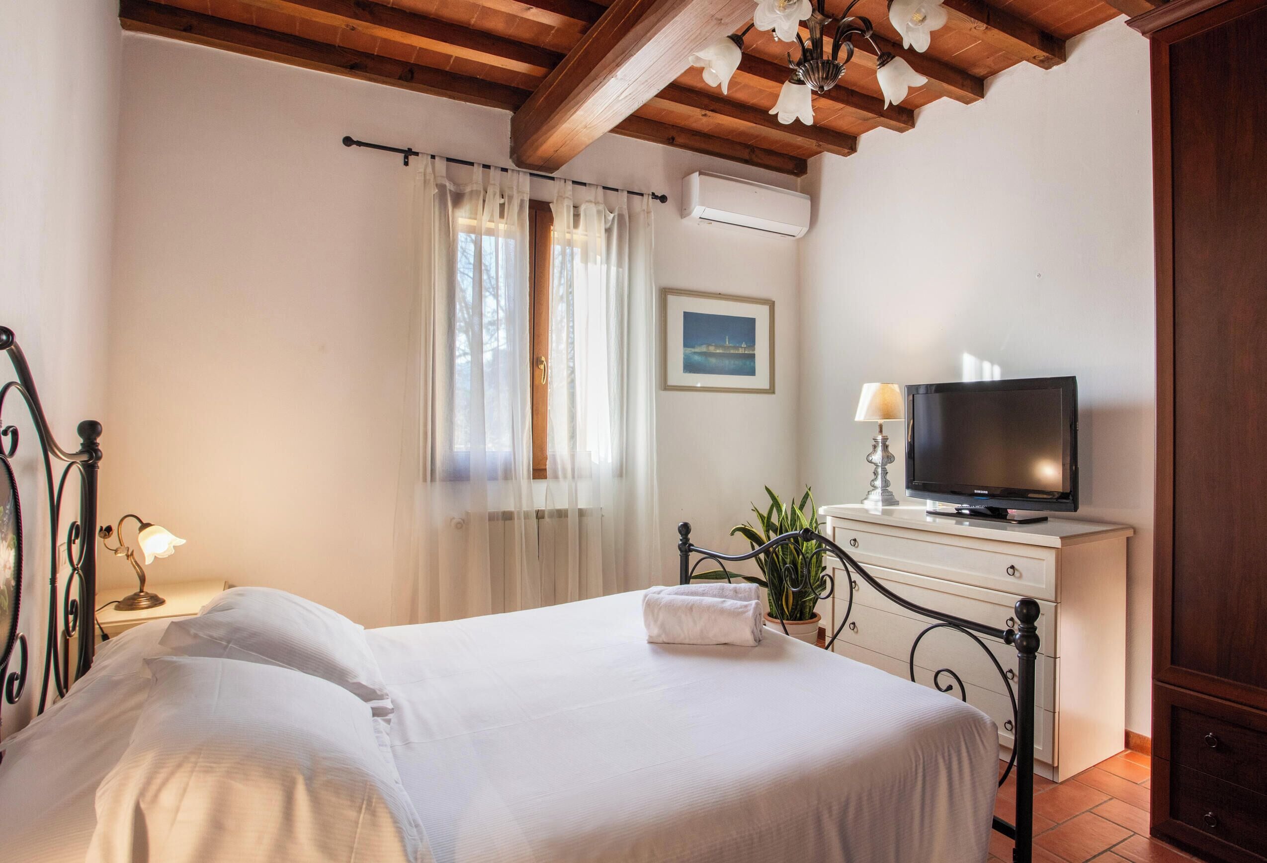 A bedroom with a bed, a television on a dresser, a window with curtains, and a picture on the wall.