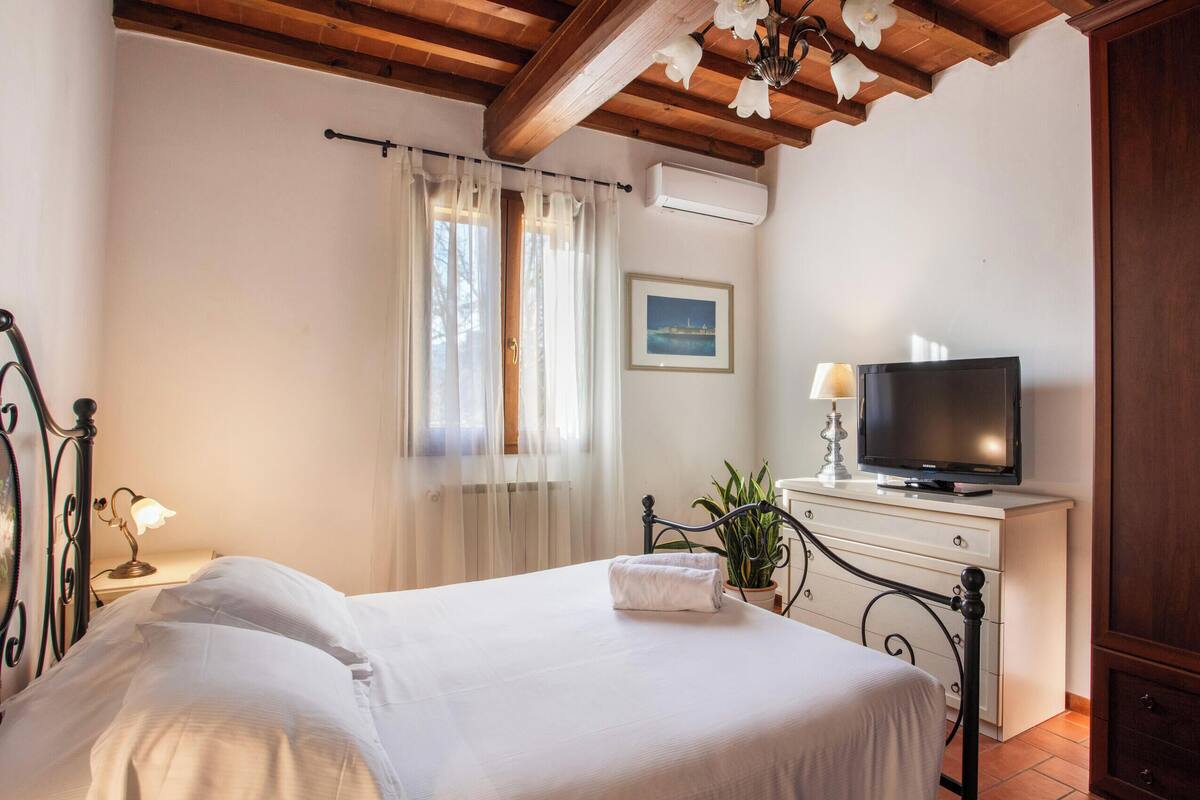 Un dormitorio con cama, televisor sobre una cómoda, ventana con cortinas e imagen en la pared.