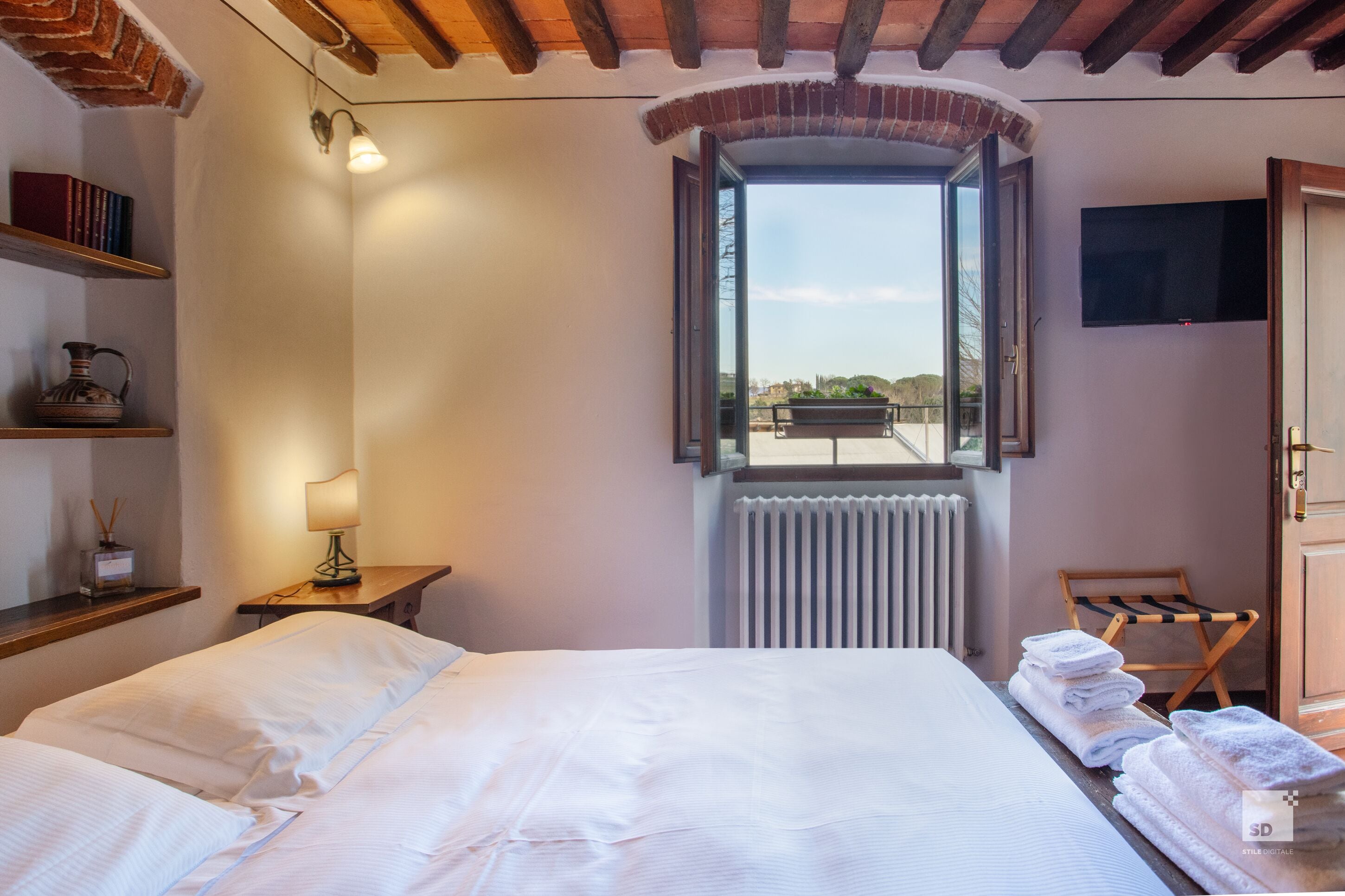 A hotel room with a bed, a window, a radiator, a TV, and a shelf with books and a lamp.