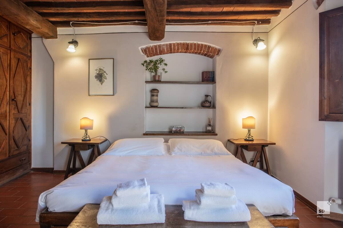 Una cama grande con ropa de cama blanca, dos mesitas de noche con lámparas, una estantería con objetos decorativos y un armario de madera.