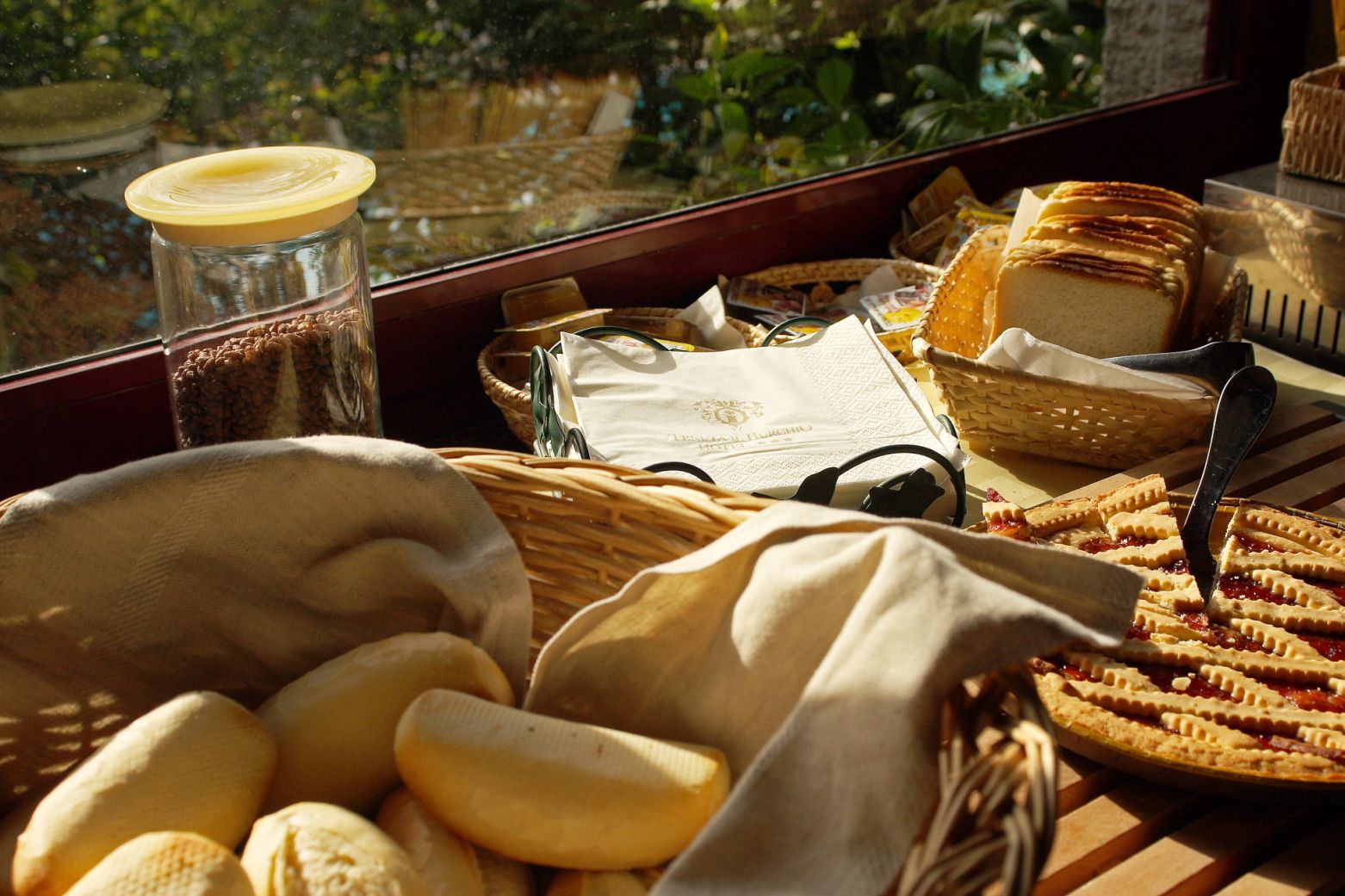 Un buffet de petit-déjeuner avec du pain, des viennoiseries et une tarte, disposé sur une table en bois près d’une fenêtre.
