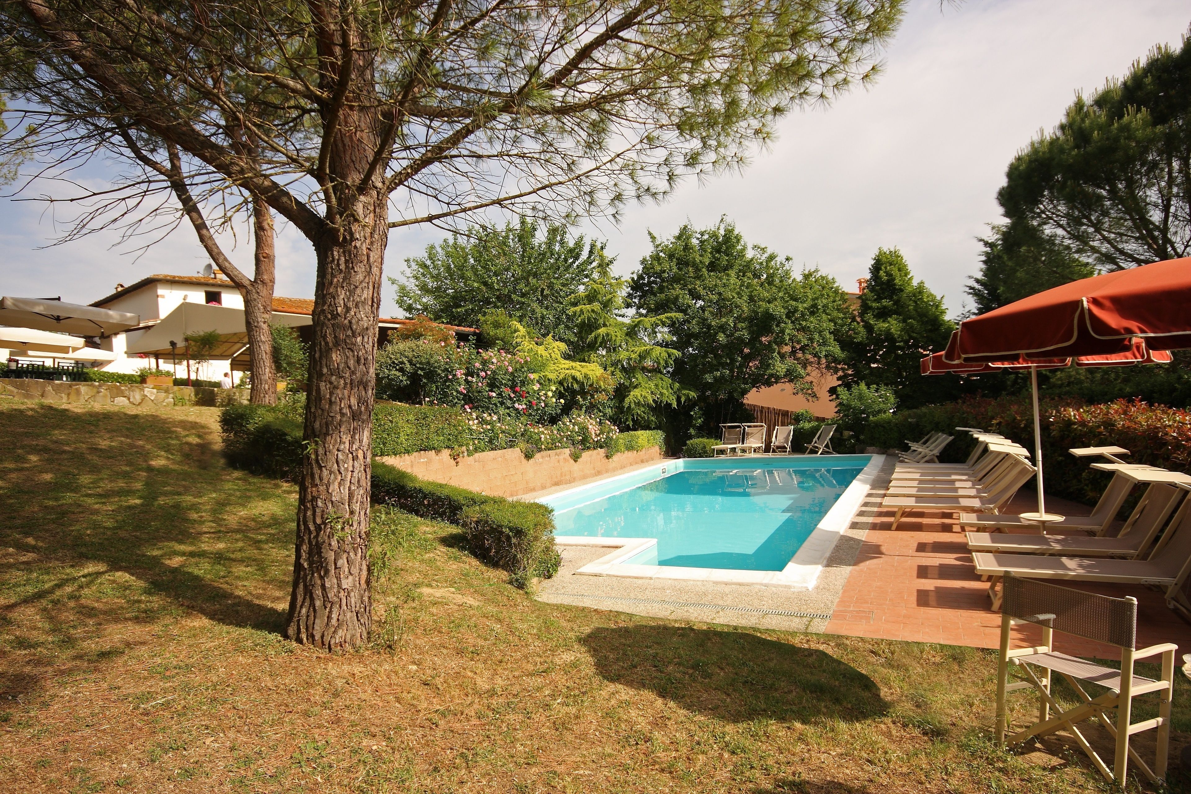 A swimming pool with lounge chairs and a red umbrella, surrounded by trees and a brick patio.
