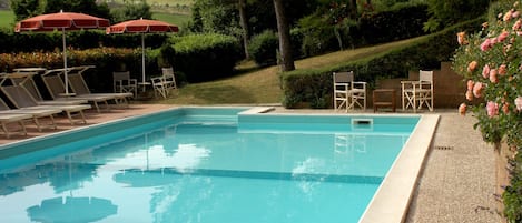 Uma piscina retangular com água azul cristalina, rodeada por espreguiçadeiras e guarda-sóis vermelhos.