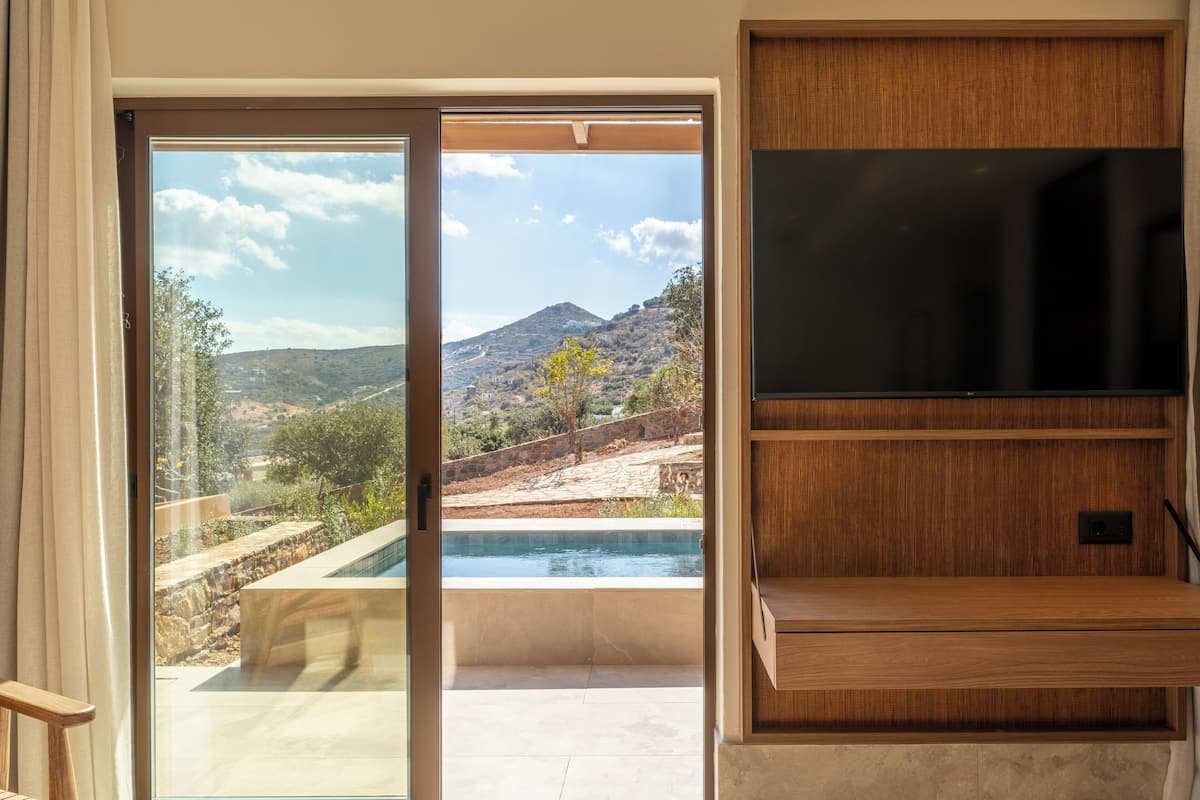 A room with a sliding glass door leading to a pool area, a flat-screen TV mounted on a wooden shelf, and a view of a mountainous landscape.