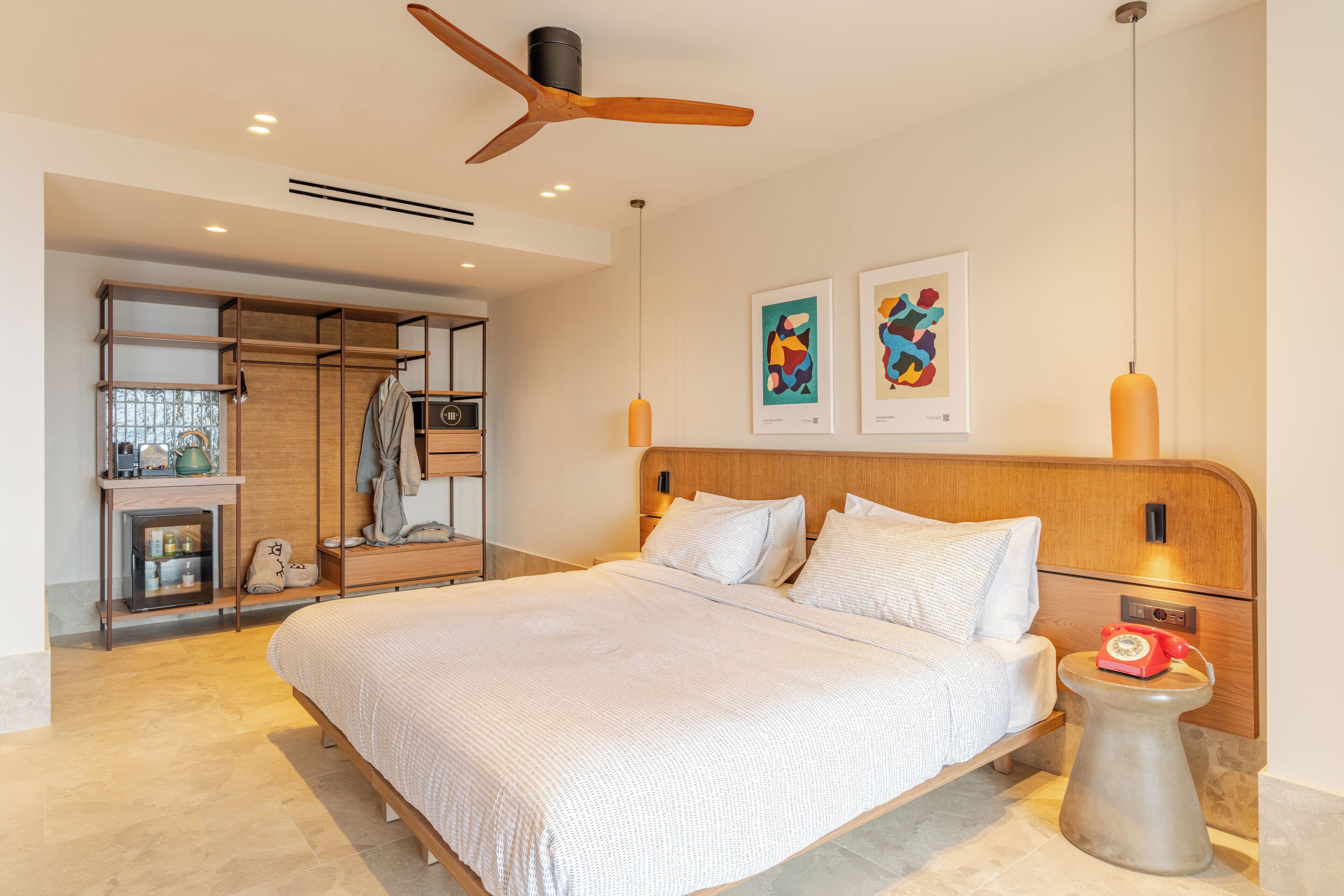 Un dormitorio moderno con una cama grande, cabecera de madera, ventilador de techo, mesita de noche con teléfono rojo y una obra de arte en la pared.