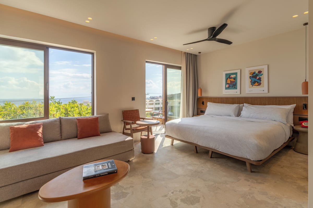 A modern hotel room with a large bed, a sofa, a small table, and a balcony with a view.