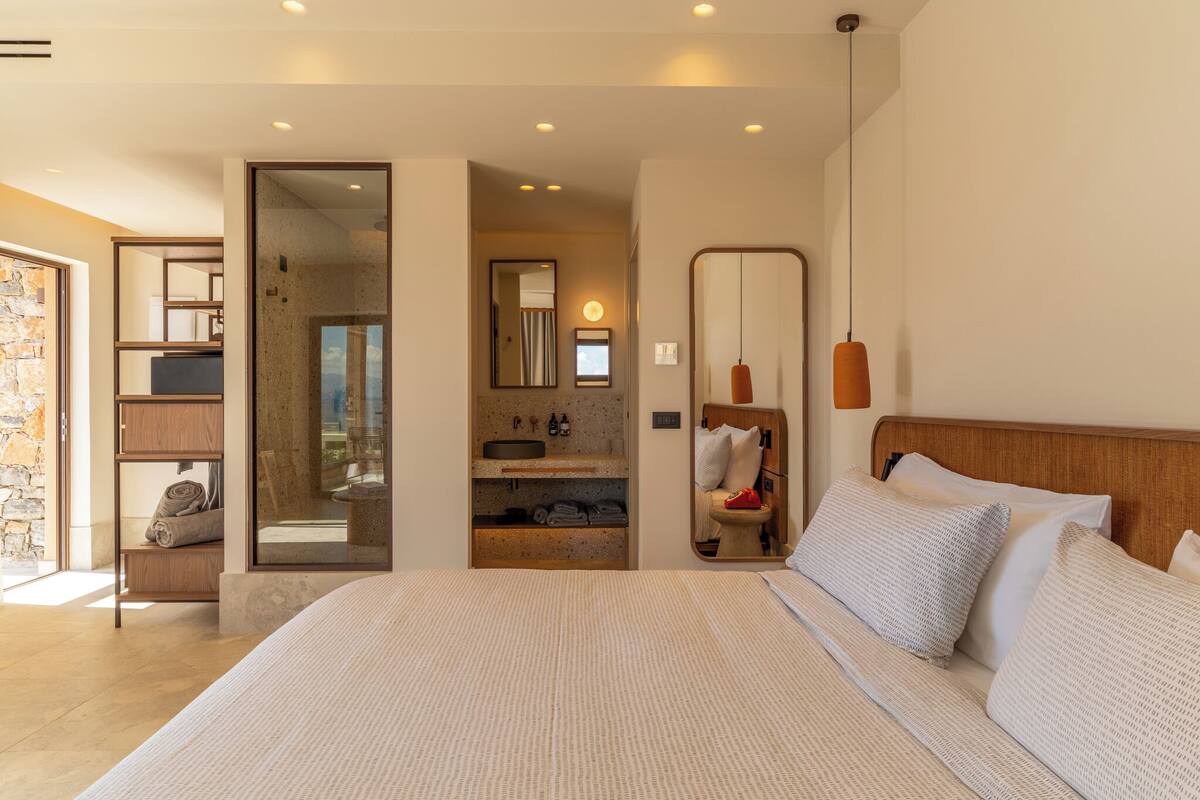 A modern hotel room with a large bed, a bathroom with a mirror, and a stone wall.