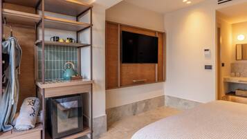 A modern hotel room with a large bed, a built-in TV, a wooden shelf with towels, and a small kitchen area.